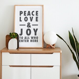 May include: A framed print with the words "PEACE LOVE & JOY TO ALL WHO ENTER HERE" in black lettering. A wooden planter with a succulent and a white sphere lamp sit on a white and wood-tone chest of drawers.