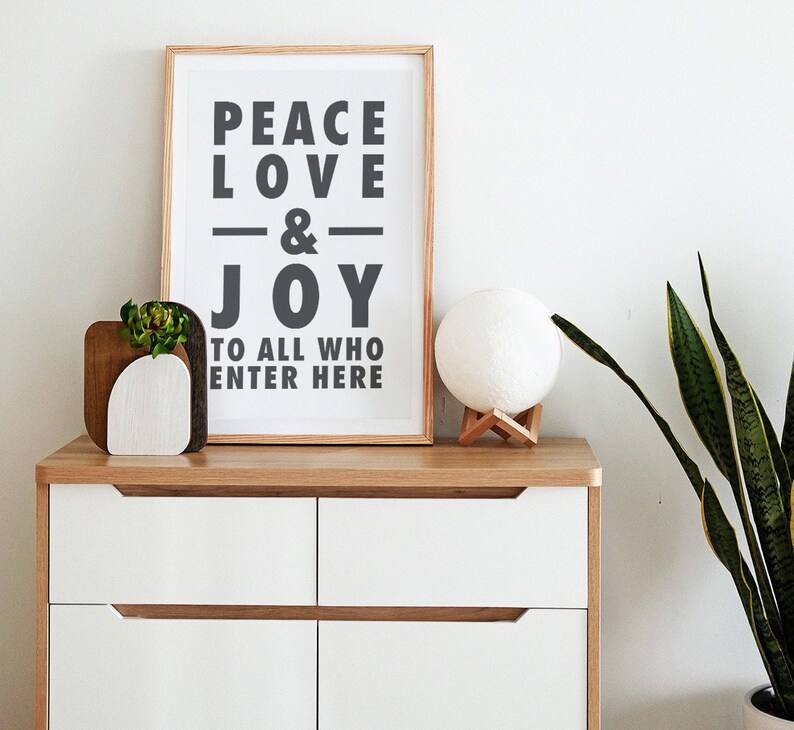 May include: A framed print with the words "PEACE LOVE & JOY TO ALL WHO ENTER HERE" in black lettering. A wooden planter with a succulent and a white sphere lamp sit on a white and wood-tone chest of drawers.