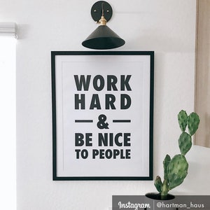 Puede incluir: Impresi&oacute;n enmarcada en blanco y negro con el texto "Work Hard & Be Nice To People" colgada en una pared blanca. Una peque&ntilde;a planta de cactus est&aacute; en una maceta negra en la esquina inferior derecha de la imagen.
