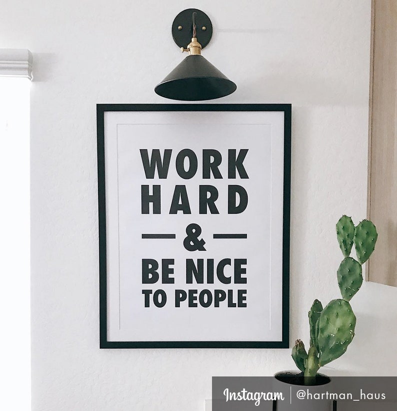 Puede incluir: Impresi&oacute;n enmarcada en blanco y negro con el texto "Work Hard & Be Nice To People" colgada en una pared blanca. Una peque&ntilde;a planta de cactus est&aacute; en una maceta negra en la esquina inferior derecha de la imagen.