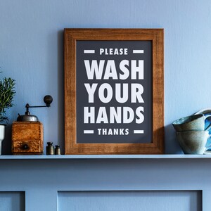 Puede incluir: Un letrero enmarcado con el texto "PLEASE WASH YOUR HANDS THANKS" en blanco sobre un fondo gris oscuro. Un molinillo de caf&eacute; de madera, pesas peque&ntilde;as y dos tazas verde azulado est&aacute;n en una estanter&iacute;a azul.