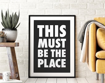 Op de afbeelding: Zwart-witte geprinte afbeelding in een zwarte lijst met de tekst "THIS MUST BE THE PLACE" in witte, vetgedrukte letters op een zwarte achtergrond.