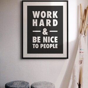 Puede incluir: Un p&oacute;ster motivacional enmarcado en blanco y negro con el texto "Trabaja duro y s&eacute; amable con la gente". El p&oacute;ster est&aacute; colgado en una pared blanca con dos taburetes tejidos grises en primer plano.