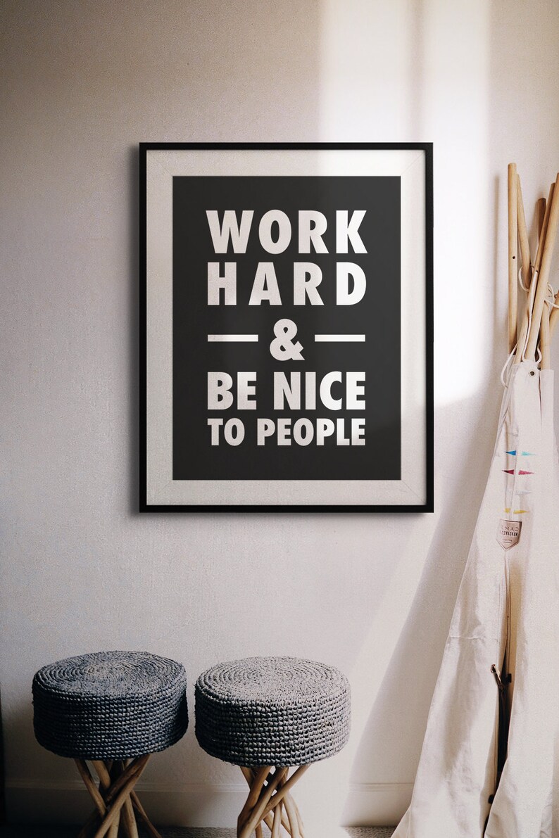Puede incluir: Un p&oacute;ster motivacional enmarcado en blanco y negro con el texto "Trabaja duro y s&eacute; amable con la gente". El p&oacute;ster est&aacute; colgado en una pared blanca con dos taburetes tejidos grises en primer plano.