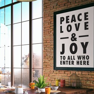 May include: A framed poster with the words "PEACE LOVE & JOY TO ALL WHO ENTER HERE" hangs on a brick wall. The room features a large window, a wooden table, and decorative items.