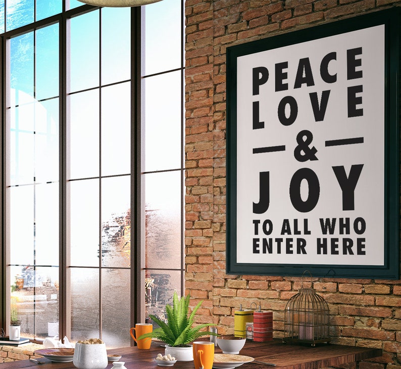 May include: A framed poster with the words "PEACE LOVE & JOY TO ALL WHO ENTER HERE" hangs on a brick wall. The room features a large window, a wooden table, and decorative items.