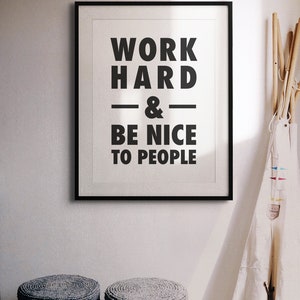 Puede incluir: Impresi&oacute;n enmarcada en blanco y negro con el texto "Work Hard & Be Nice To People" colgada en una pared blanca. Dos taburetes tejidos grises est&aacute;n en primer plano.