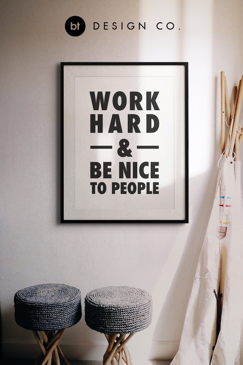 Puede incluir: Impresi&oacute;n enmarcada en blanco y negro con el texto "Work Hard & Be Nice To People" colgada en una pared blanca. Dos taburetes tejidos grises est&aacute;n en primer plano.