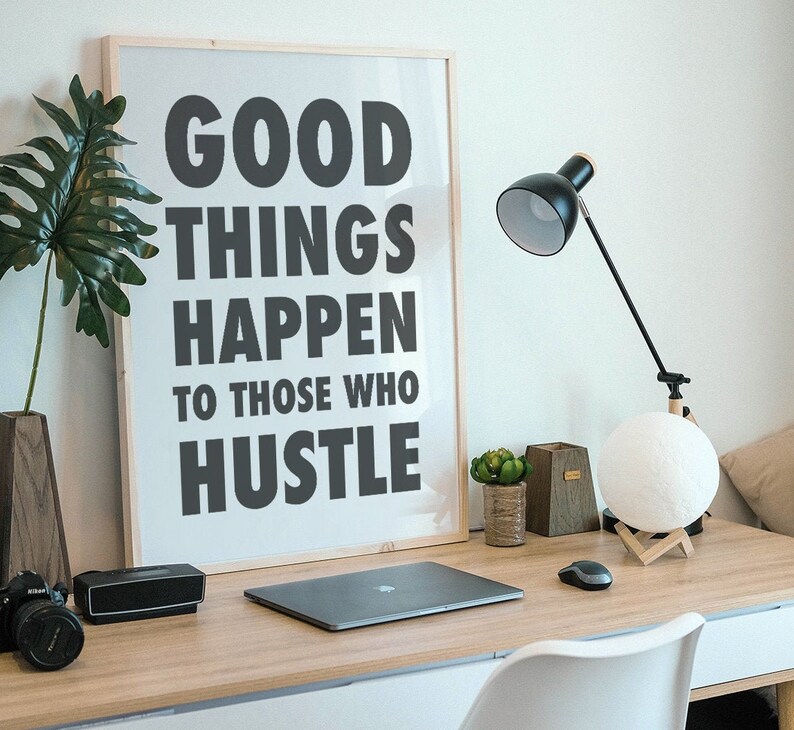 May include: A home office desk with a framed print that reads "GOOD THINGS HAPPEN TO THOSE WHO HUSTLE." The desk also features a laptop, a black desk lamp, a moon-shaped lamp, a camera, and a small succulent.