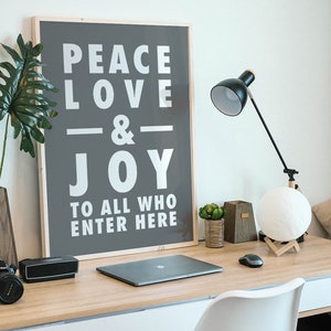 May include: A framed poster with the text "Peace Love & Joy To All Who Enter Here" on a gray background. The poster is hanging on a white wall above a desk with a laptop, a lamp, and a plant.