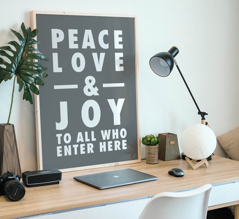 May include: A framed poster with the text "Peace Love & Joy To All Who Enter Here" on a gray background. The poster is hanging on a white wall above a desk with a laptop, a lamp, and a plant.