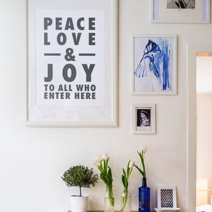 May include: A collection of framed art and decorative items on a white wall. A large framed print reads "PEACE LOVE & JOY TO ALL WHO ENTER HERE." Other framed pieces include a blue watercolour and a portrait. White tulips in glass vases and a small potted plant are also present.