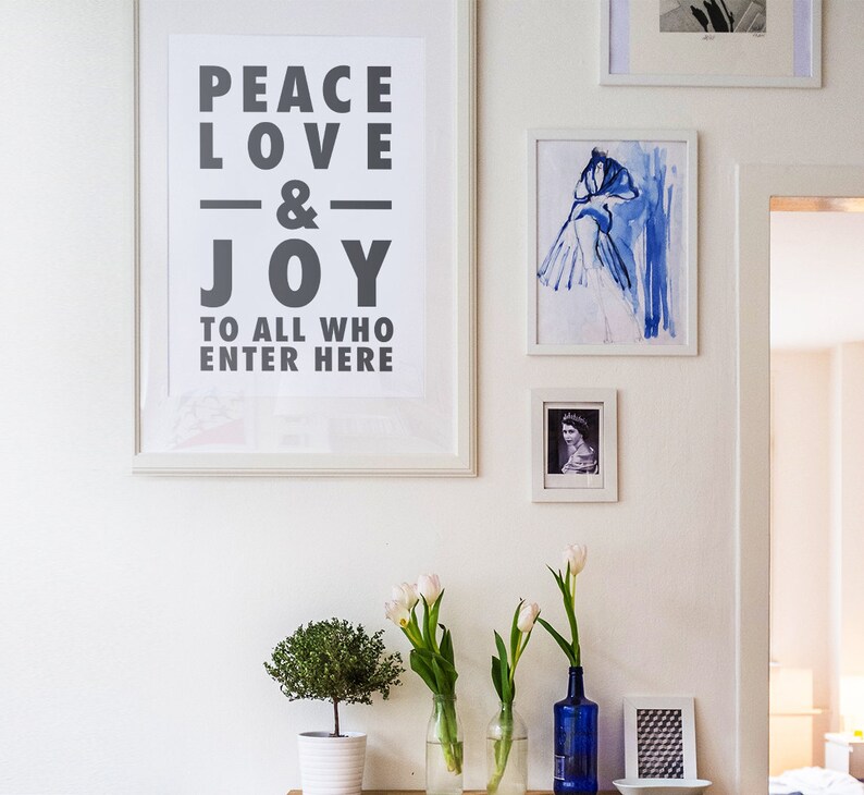 May include: A collection of framed art and decorative items on a white wall. A large framed print reads "PEACE LOVE & JOY TO ALL WHO ENTER HERE." Other framed pieces include a blue watercolour and a portrait. White tulips in glass vases and a small potted plant are also present.