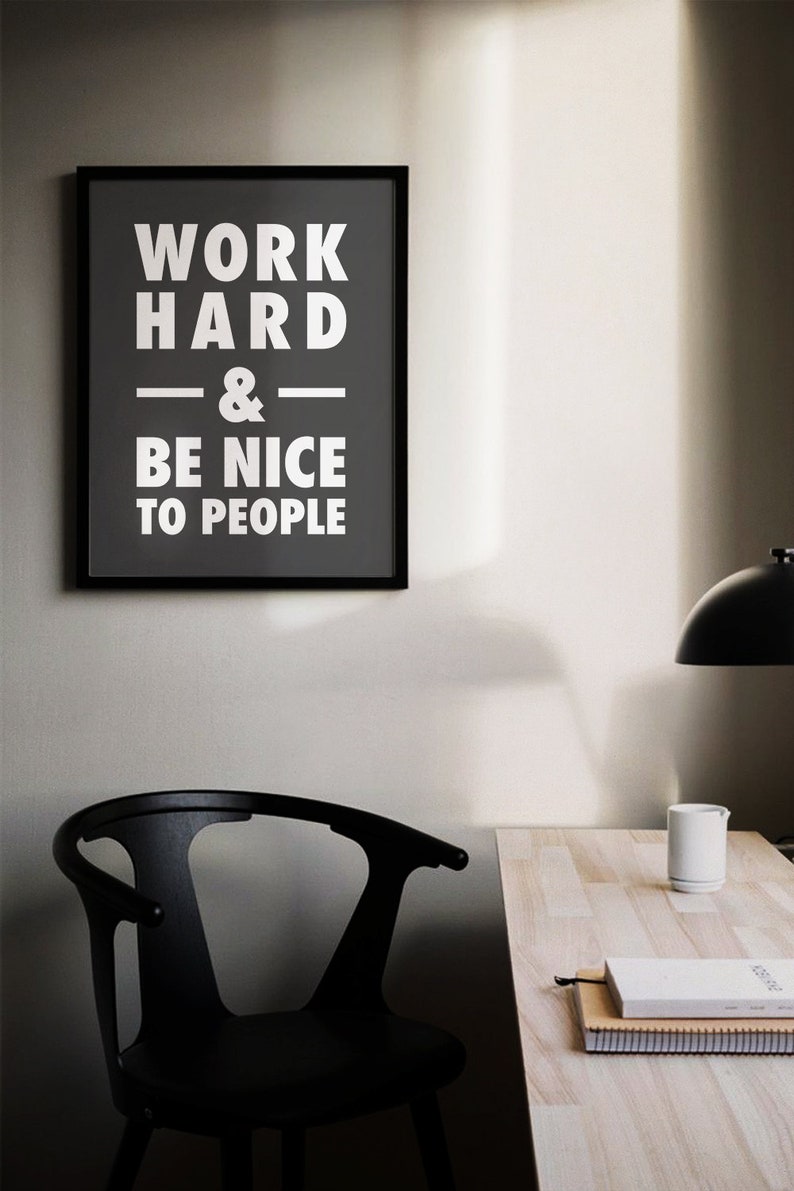 Puede incluir: Un p&oacute;ster motivacional en blanco y negro con el texto "Trabaja duro y s&eacute; amable con la gente" colgado en una pared gris sobre una silla negra y un escritorio de madera.