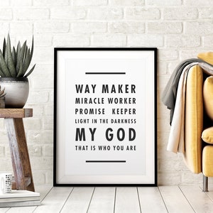 May include: A framed print with the words "WAY MAKER, MIRACLE WORKER, PROMISE KEEPER, LIGHT IN THE DARKNESS, MY GOD, THAT IS WHO YOU ARE" in black text on a white background. The frame is black. The print is displayed near a wooden stool with plants and a yellow sofa.