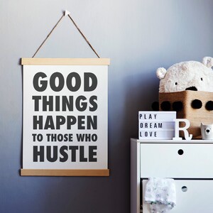 May include: A white poster with the text "GOOD THINGS HAPPEN TO THOSE WHO HUSTLE" hangs from a wooden frame. A white dresser holds a bear-shaped basket, a light-up sign, and a ceramic bear cup. The wall is painted a muted blue.