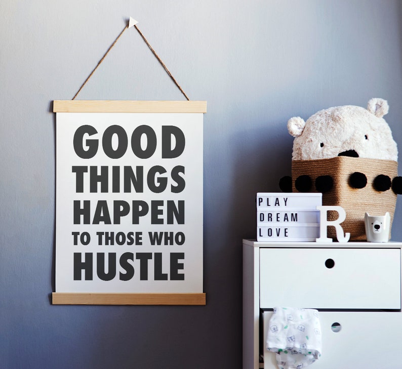 May include: A white poster with the text "GOOD THINGS HAPPEN TO THOSE WHO HUSTLE" hangs from a wooden frame. A white dresser holds a bear-shaped basket, a light-up sign, and a ceramic bear cup. The wall is painted a muted blue.