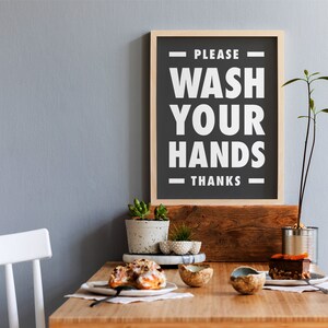 Puede incluir: Un letrero enmarcado con el texto "PLEASE WASH YOUR HANDS THANKS" en blanco sobre un fondo negro. El letrero se muestra encima de una mesa de madera con peque&ntilde;as plantas en macetas, pasteles y una rebanada de pastel. El fondo es una pared gris claro.