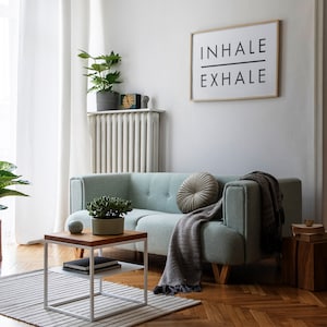 Op de afbeelding: Een lichtgroene bank met een grijze deken en een rond kussen. De bank staat in een woonkamer met een witte radiator, een witte salontafel met twee planken en een ingelijst printje met de tekst "INHALE EXHALE".