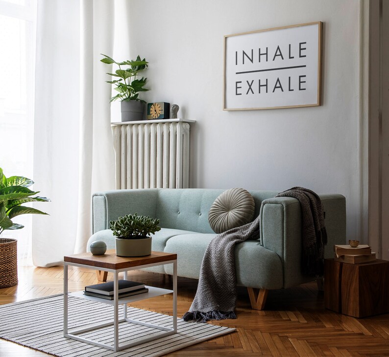 Op de afbeelding: Een lichtgroene bank met een grijze deken en een rond kussen. De bank staat in een woonkamer met een witte radiator, een witte salontafel met twee planken en een ingelijst printje met de tekst "INHALE EXHALE".