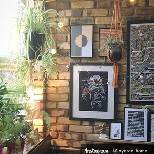 May include: A brick wall with a variety of framed artwork, including a black and white botanical print, a geometric print, and a colorful map. There are also several plants hanging from the wall, including a spider plant and a hanging plant in a macrame planter. The wall is decorated with string lights and the text "Instagram | @layered.home" is visible at the bottom of the image.