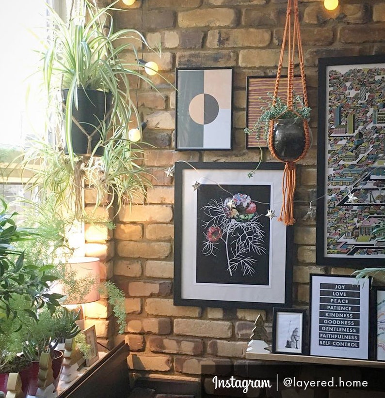 May include: A brick wall with a variety of framed artwork, including a black and white botanical print, a geometric print, and a colorful map. There are also several plants hanging from the wall, including a spider plant and a hanging plant in a macrame planter. The wall is decorated with string lights and the text "Instagram | @layered.home" is visible at the bottom of the image.