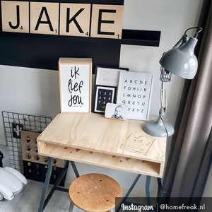May include: A light wood desk with a gray metal frame and a shelf underneath. There is a gray lamp with a cage shade on the desk. There are also several framed prints on the wall behind the desk, including one with the word "JAKE" in black letters on white blocks.