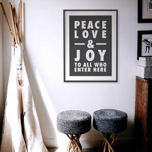 May include: A black and white framed print with the text "Peace Love & Joy To All Who Enter Here" hanging on a white wall. Two gray woven stools with wooden legs are in the foreground.