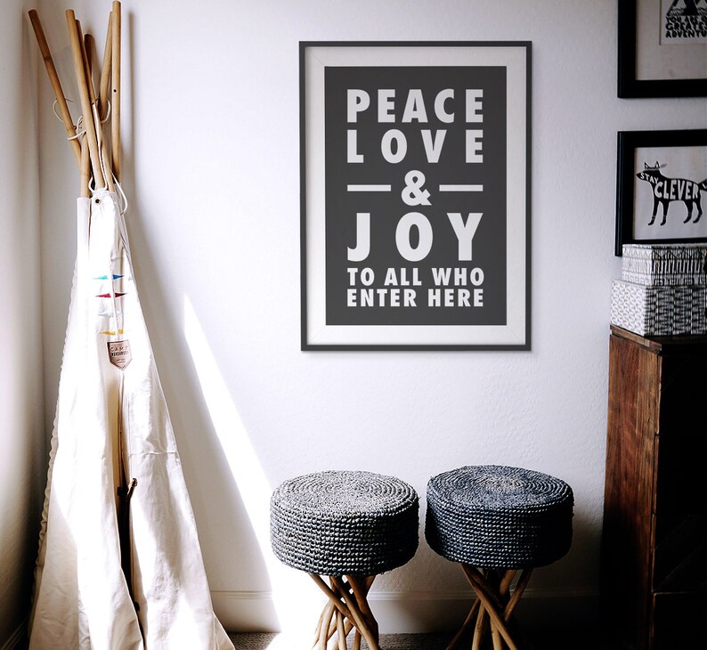 May include: A black and white framed print with the text "Peace Love & Joy To All Who Enter Here" hanging on a white wall. Two gray woven stools with wooden legs are in the foreground.