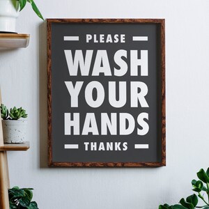 Puede incluir: Un letrero enmarcado con un fondo gris oscuro y texto blanco que dice "PLEASE WASH YOUR HANDS THANKS". El marco es de madera con un acabado marr&oacute;n oscuro. El letrero est&aacute; colgado en una pared blanca.