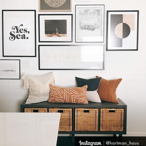 May include: A black bench with three woven baskets and a white table in front of it. The bench is decorated with a variety of pillows, including a white fringed pillow, a brown and white patterned pillow, a dark brown pillow, and a light brown pillow. The wall behind the bench is decorated with black framed artwork, including a print that says "Yes, Sea."