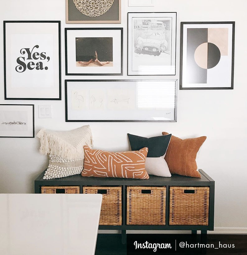 May include: A black bench with three woven baskets and a white table in front of it. The bench is decorated with a variety of pillows, including a white fringed pillow, a brown and white patterned pillow, a dark brown pillow, and a light brown pillow. The wall behind the bench is decorated with black framed artwork, including a print that says "Yes, Sea."