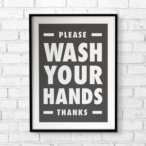 Puede incluir: Un cartel enmarcado con un borde negro y un paspart&uacute; blanco. El cartel tiene un fondo gris oscuro con las palabras "PLEASE WASH YOUR HANDS THANKS" en letras may&uacute;sculas blancas grandes. El cartel est&aacute; colgado en una pared de ladrillos blancos.