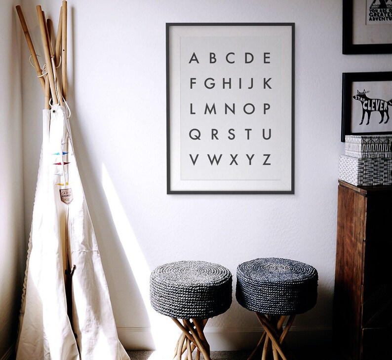 May include: A black framed print with the alphabet in white letters on a white background. Two gray woven stools with wooden legs are in the foreground.