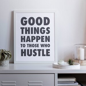 May include: A framed print with the text "GOOD THINGS HAPPEN TO THOSE WHO HUSTLE" in dark gray. The print is displayed on a gray shelf with a small succulent in a concrete pot, a decorative tray, and a glass candle holder.