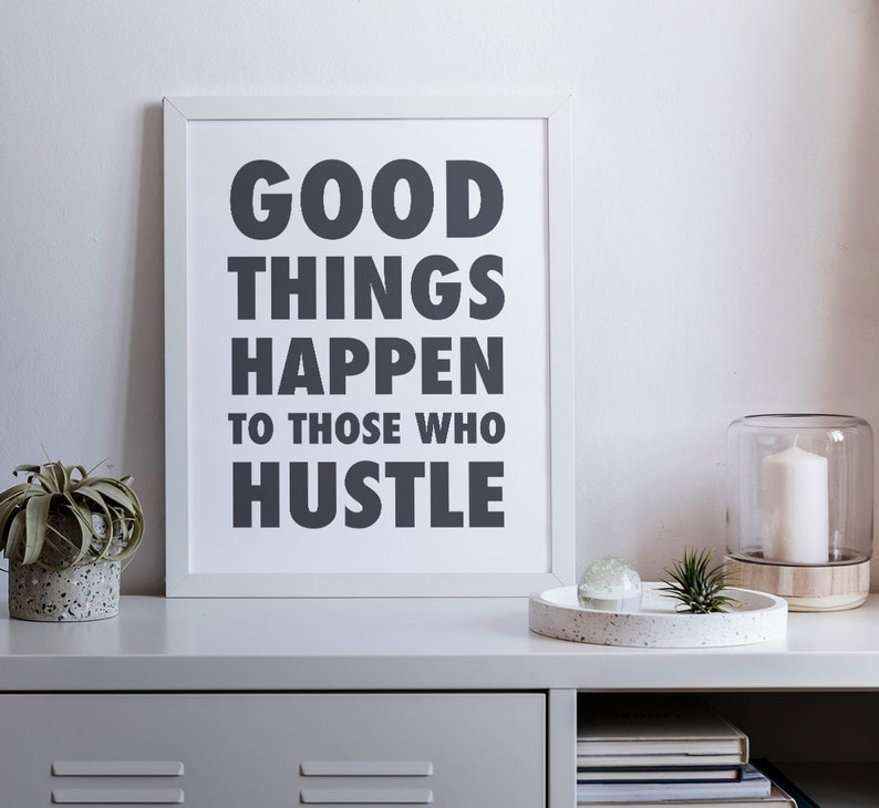 May include: A framed print with the text "GOOD THINGS HAPPEN TO THOSE WHO HUSTLE" in dark gray. The print is displayed on a gray shelf with a small succulent in a concrete pot, a decorative tray, and a glass candle holder.