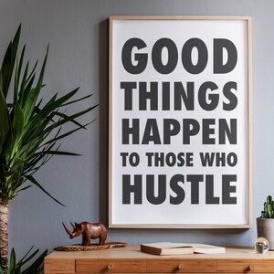 May include: A framed print with the text "GOOD THINGS HAPPEN TO THOSE WHO HUSTLE" in bold, black letters. The print is displayed on a wooden desk with a wooden rhino figurine, a book, and a potted plant.
