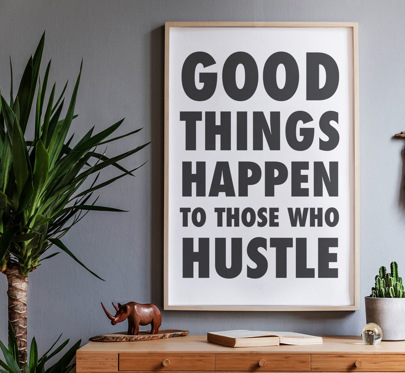 May include: A framed print with the text "GOOD THINGS HAPPEN TO THOSE WHO HUSTLE" in bold, black letters. The print is displayed on a wooden desk with a wooden rhino figurine, a book, and a potted plant.