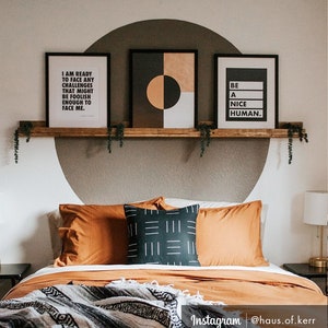 May include: A bedroom with a wooden shelf above the bed. The shelf has three framed prints on it. The print on the left says "I am ready to face any challenges that might be foolish enough to face me." The middle print is a geometric design with black, white, and orange shapes. The print on the right says "Be a nice human." The bed has a brown headboard and a brown and white blanket on it.