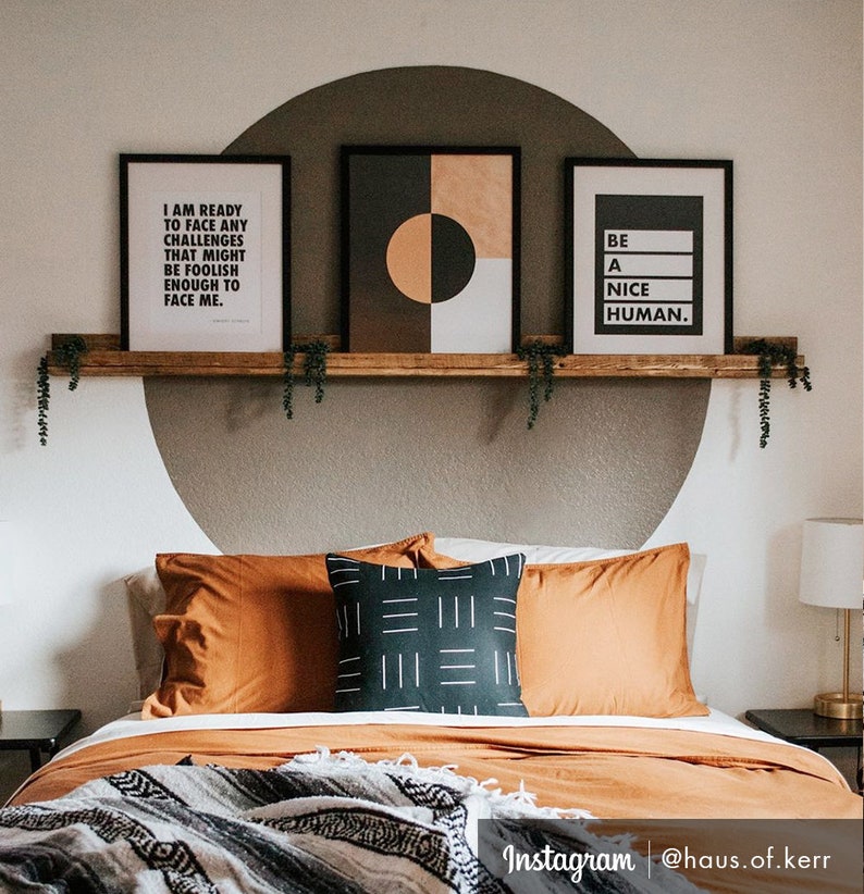May include: A bedroom with a wooden shelf above the bed. The shelf has three framed prints on it. The print on the left says "I am ready to face any challenges that might be foolish enough to face me." The middle print is a geometric design with black, white, and orange shapes. The print on the right says "Be a nice human." The bed has a brown headboard and a brown and white blanket on it.