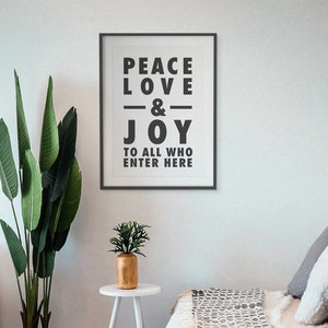 May include: A framed print with the words "PEACE LOVE & JOY TO ALL WHO ENTER HERE" in bold black letters. The print is on a white wall, with a tall green plant and a small gold vase on a white table in front of it.