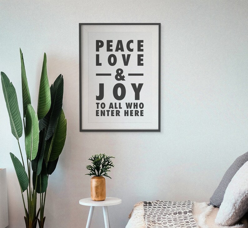 May include: A framed print with the words "PEACE LOVE & JOY TO ALL WHO ENTER HERE" in bold black letters. The print is on a white wall, with a tall green plant and a small gold vase on a white table in front of it.