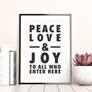 May include: A framed print with the words "PEACE LOVE & JOY TO ALL WHO ENTER HERE" in bold black letters. The print is in a black frame and is displayed on a white surface with books and a potted succulent.
