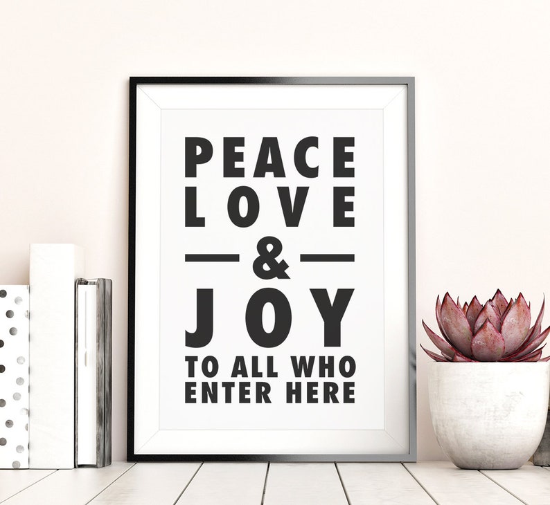 May include: A framed print with the words "PEACE LOVE & JOY TO ALL WHO ENTER HERE" in bold black letters. The print is in a black frame and is displayed on a white surface with books and a potted succulent.