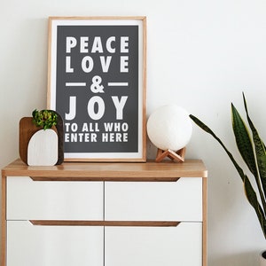 May include: A white dresser with three drawers and a wooden top. A framed print with the text "Peace Love & Joy to all who enter here" is on the top of the dresser. A white moon lamp with a wooden base is on the dresser next to the print. A small potted plant is on the dresser to the left of the print.