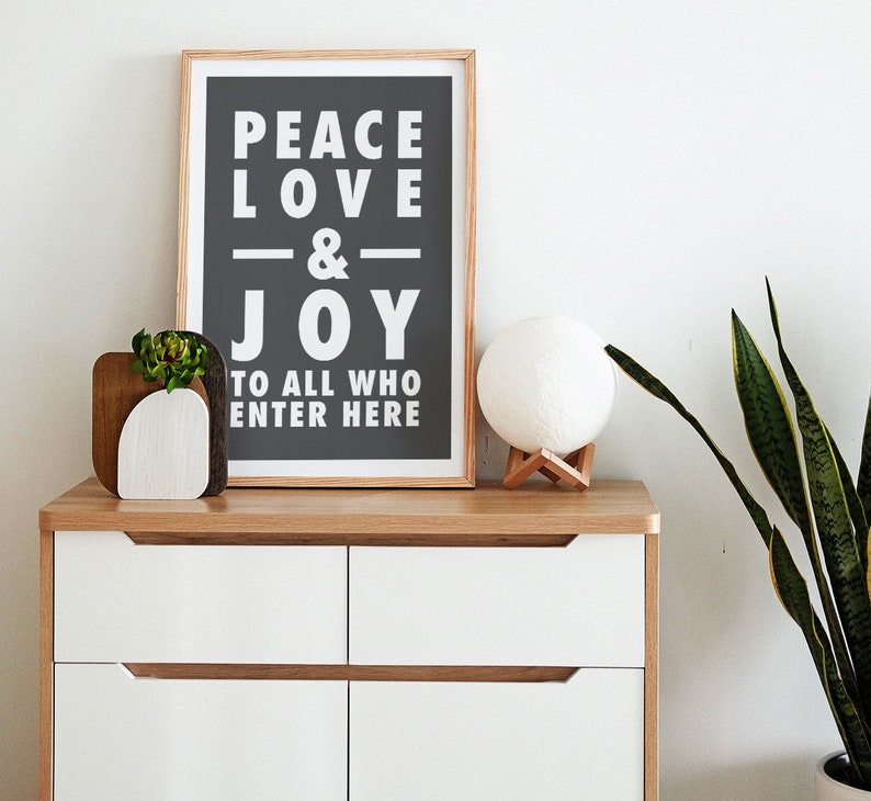 May include: A white dresser with three drawers and a wooden top. A framed print with the text "Peace Love & Joy to all who enter here" is on the top of the dresser. A white moon lamp with a wooden base is on the dresser next to the print. A small potted plant is on the dresser to the left of the print.
