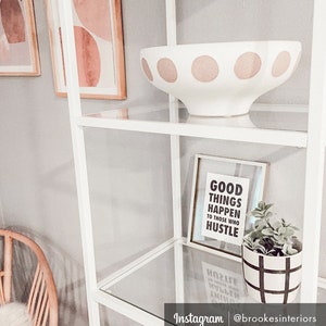 May include: White decorative bowl with large pink polka dots on a glass shelf. A framed print with the text "GOOD THINGS HAPPEN TO THOSE WHO HUSTLE" and a small potted plant are also on the shelf. A white shelving unit is in the background.