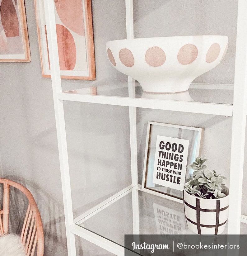 May include: White decorative bowl with large pink polka dots on a glass shelf. A framed print with the text "GOOD THINGS HAPPEN TO THOSE WHO HUSTLE" and a small potted plant are also on the shelf. A white shelving unit is in the background.