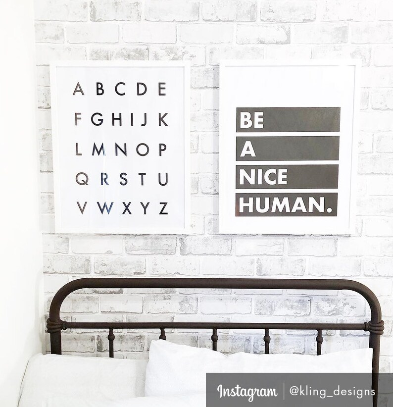 May include: Two framed prints on a white brick wall. The print on the left shows the alphabet in black letters on a white background. The print on the right has the text "BE A NICE HUMAN." in black letters on a gray background.