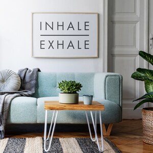 Op de afbeelding: Een lichtblauwe bank met een grijze deken en een rond grijs kussen. Een kleine houten tafel met witte haarspeldpoten staat voor de bank. Een potplant met een vetplant staat op de tafel, en een grijze keramische mok staat ernaast. Een ingelijst afdruk met de tekst "INHALE EXHALE" hangt aan de muur achter de bank.